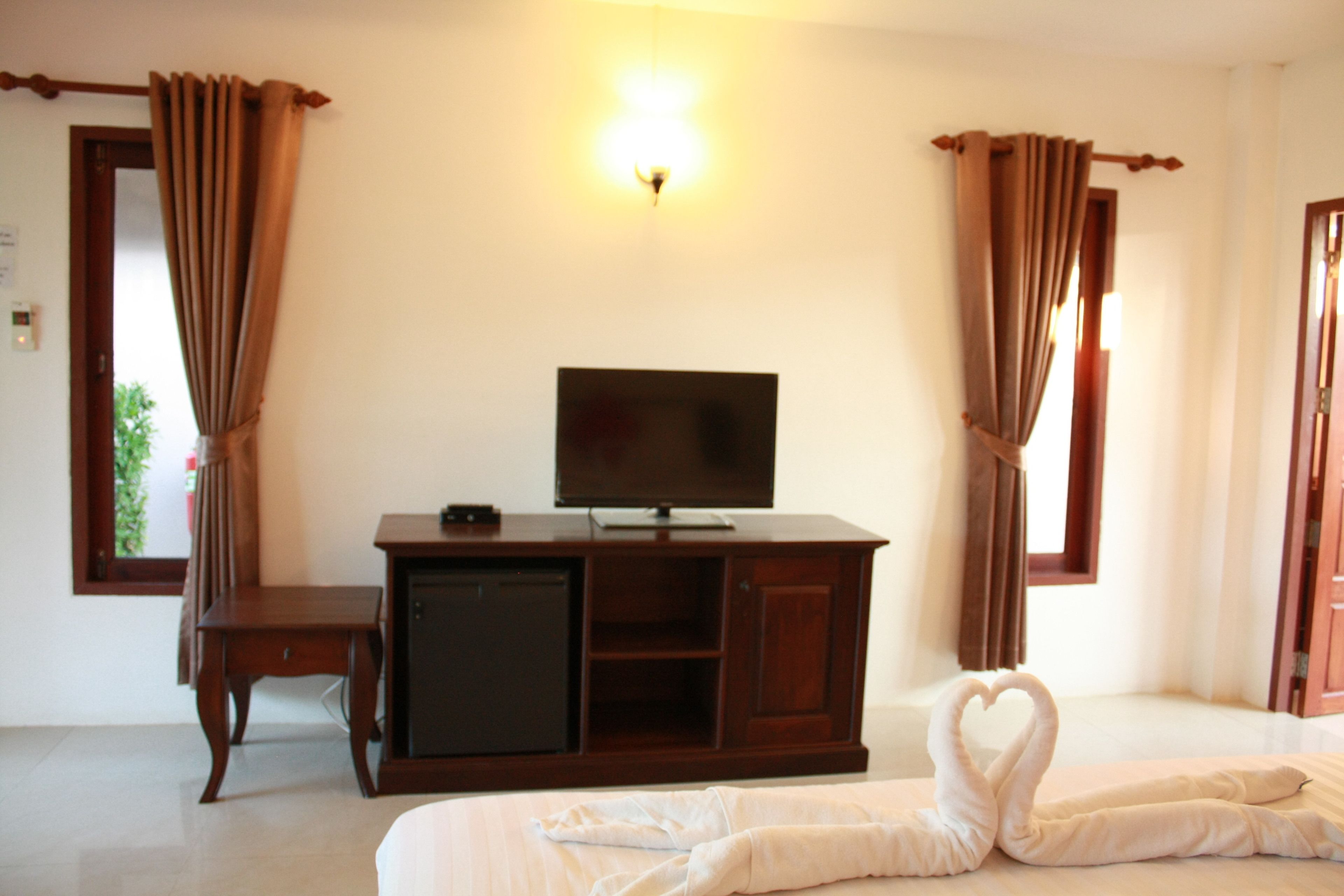 Una habitación de hotel con una televisión sobre un mueble de madera, una cama con una disposición de toallas con forma de corazón y cortinas en las ventanas.