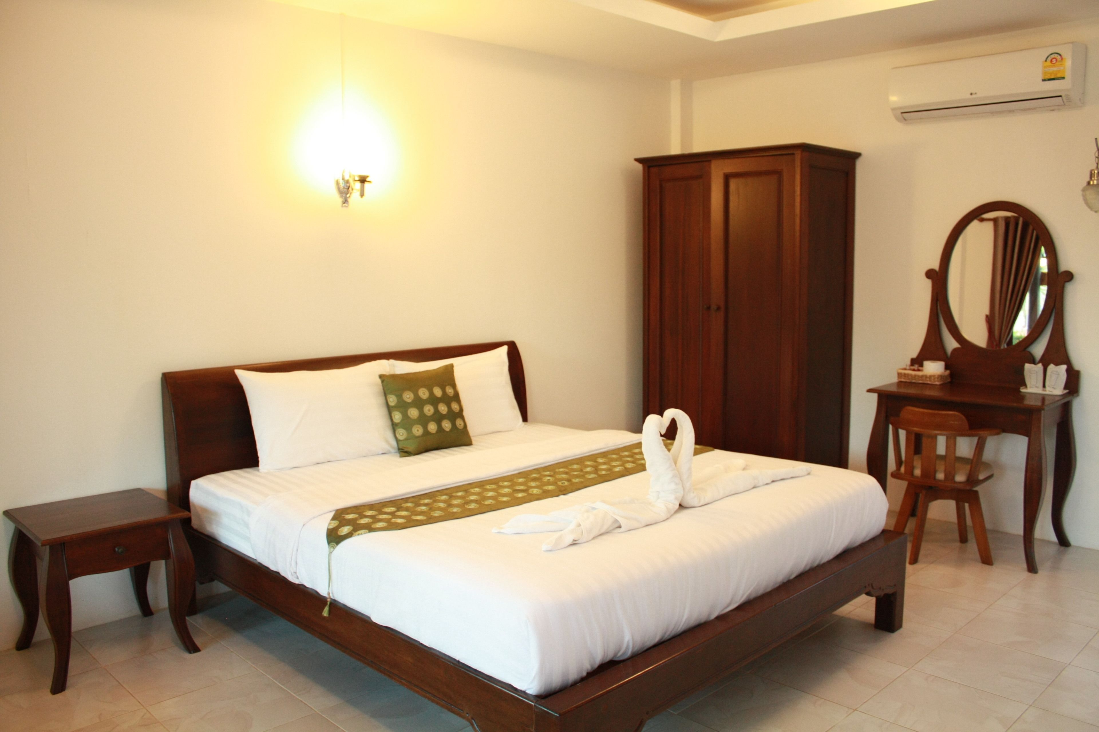 Una cama bien hecha con sábanas blancas y un almohadón verde, una mesita de noche de madera, un armario de madera y un escritorio con espejo.