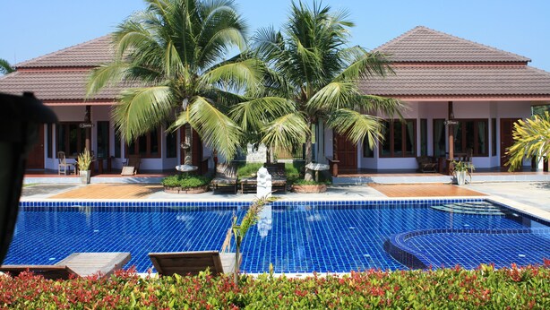 A hotel with a swimming pool, palm trees, and a tiled roof. - Waterside Resort (Pranburi)
