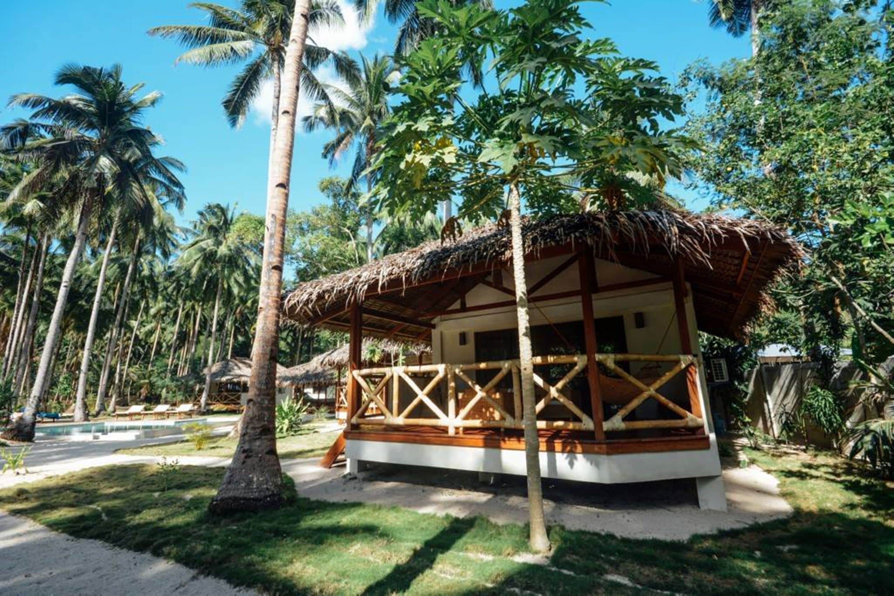 A thatched-roof bungalow with a wooden deck, surrounded by palm trees and a pool.