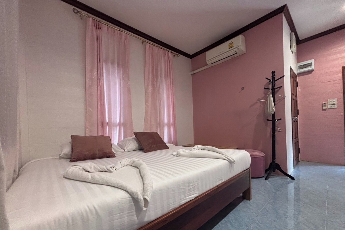 A double bed with a wooden frame, two pillows, and a white blanket. There is a wooden floor and a wall-mounted air conditioner.