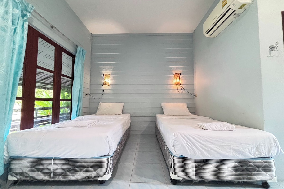 A room with two beds, a window with curtains, an air conditioning unit, and wall-mounted lights.