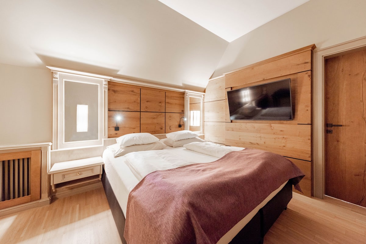 Ein Schlafzimmer mit einem Bett, einem an der Wand befestigten Fernseher und einem Fenster, das für natürliches Licht sorgt.