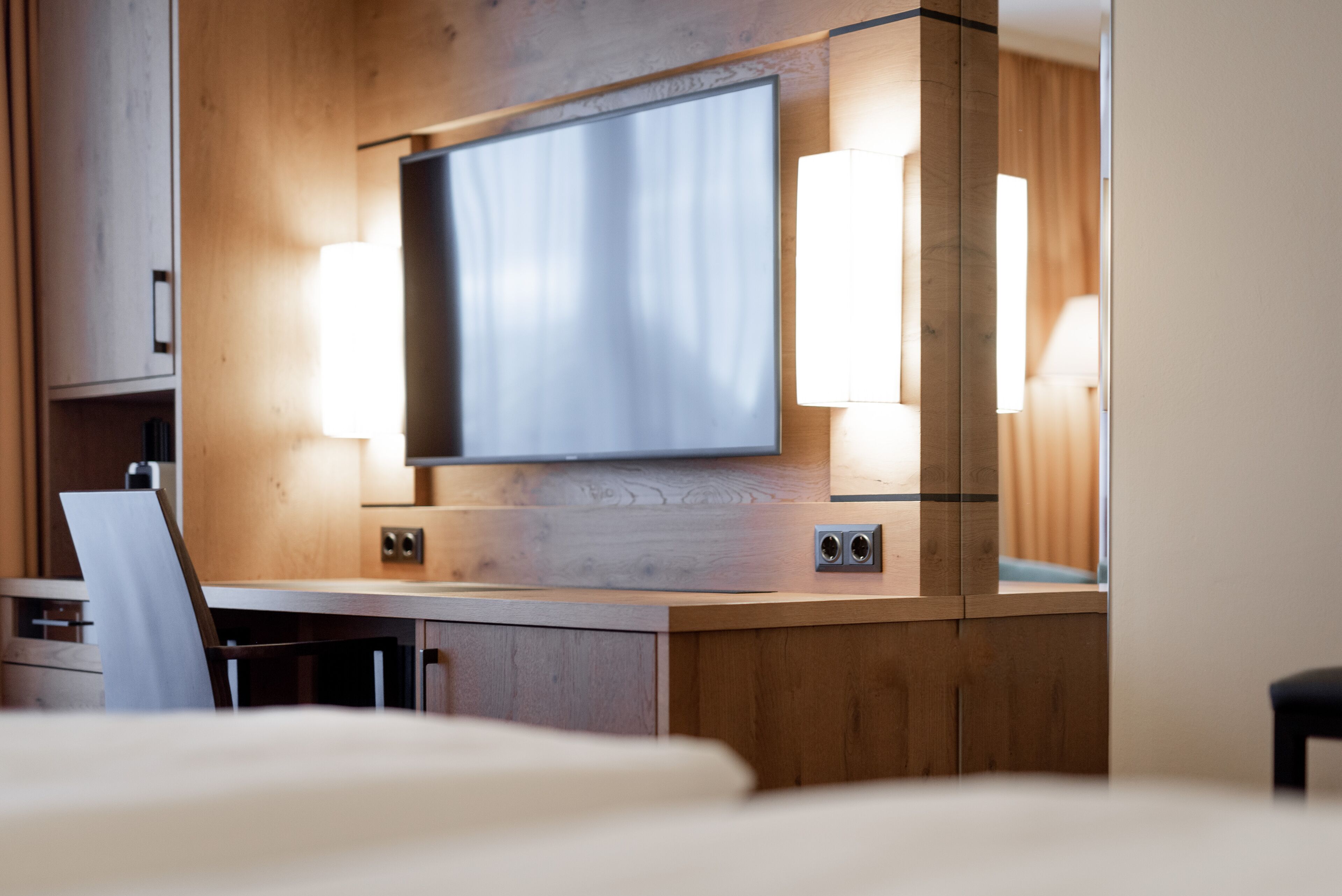 A modern hotel room with a large flat-screen TV mounted on a wooden wall, a desk with a chair, and two bedside lamps.