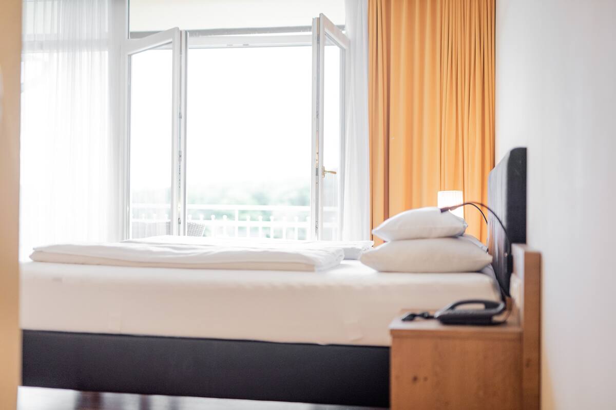 Ein ordentlich bezogenes Bett mit weißen Leinen, ein Nachttisch mit Lampe und ein Fenster mit Vorhängen.