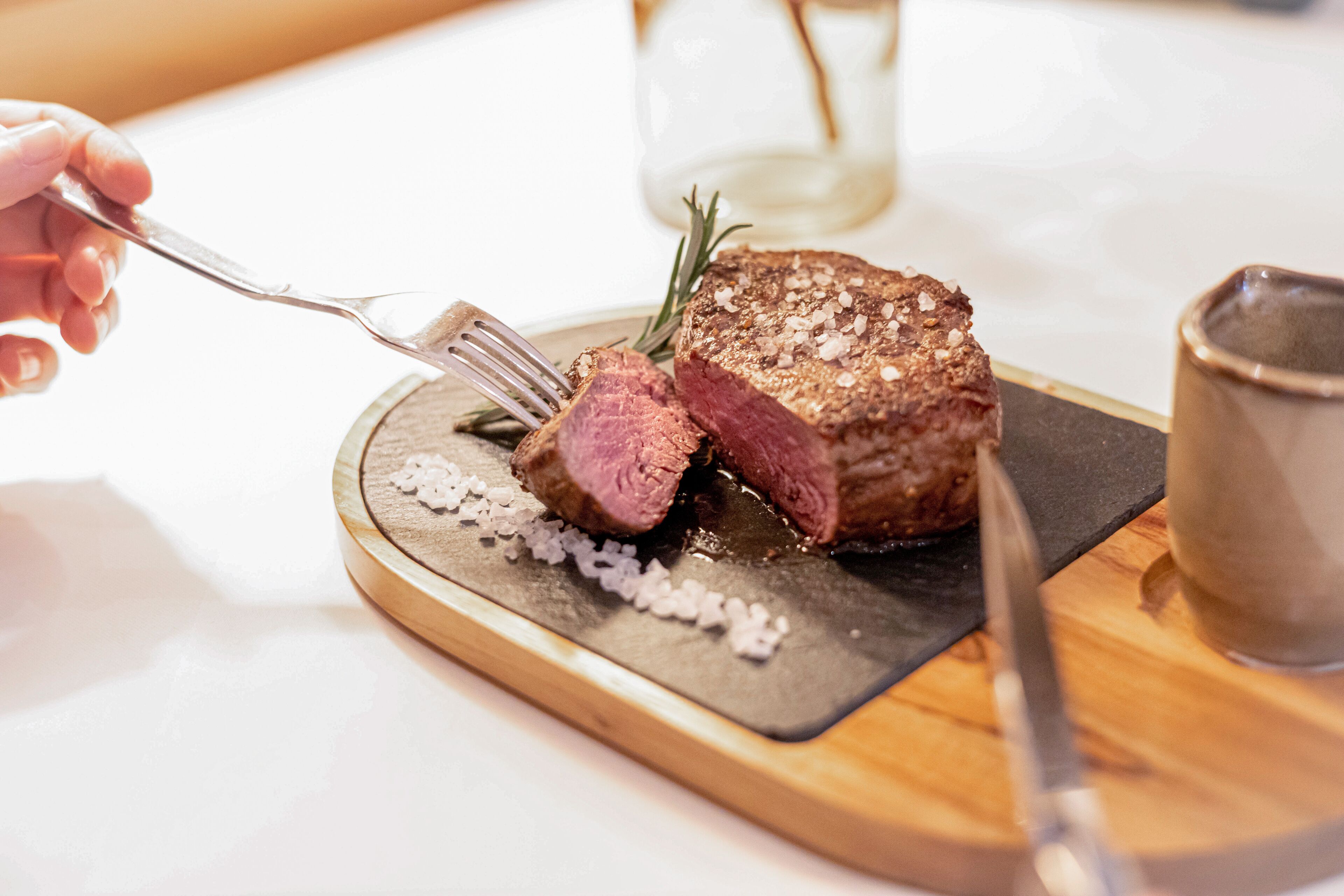 A steak with a fork and knife on a wooden board, with a glass and salt shakers in the background.