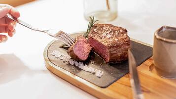 A steak with a fork and knife on a wooden board, with a glass and salt shakers in the background.