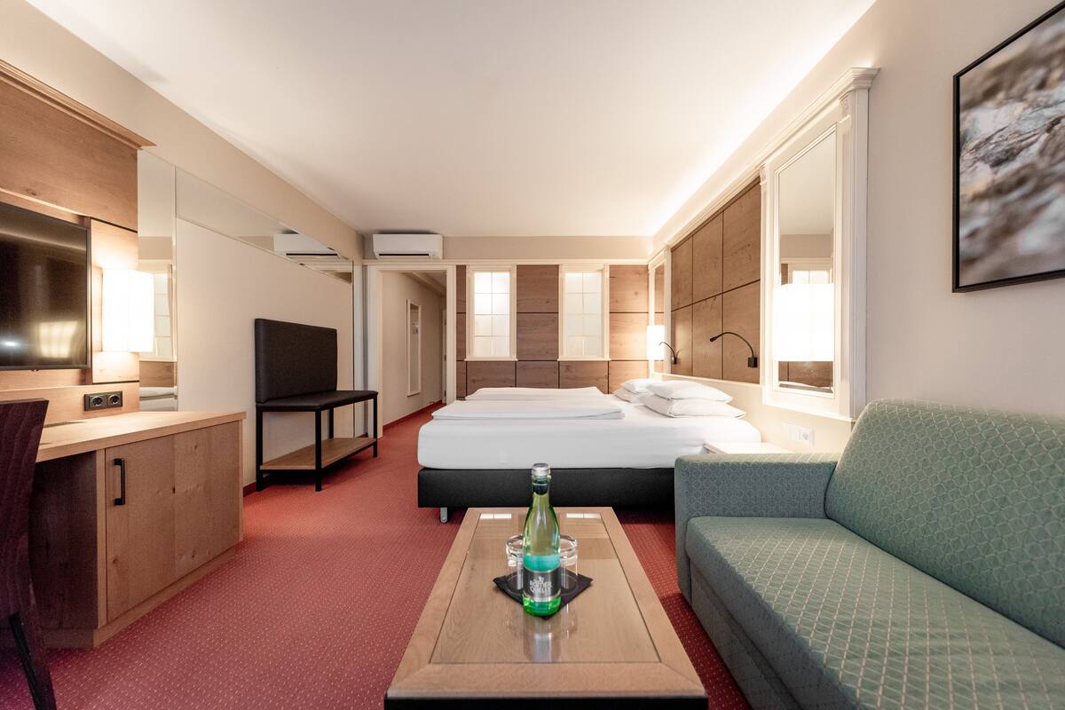 Ein Hotelzimmer mit einem Bett, einem grünen Sofa, einem Holztisch, einem Fernseher und einer Flasche auf dem Tisch.