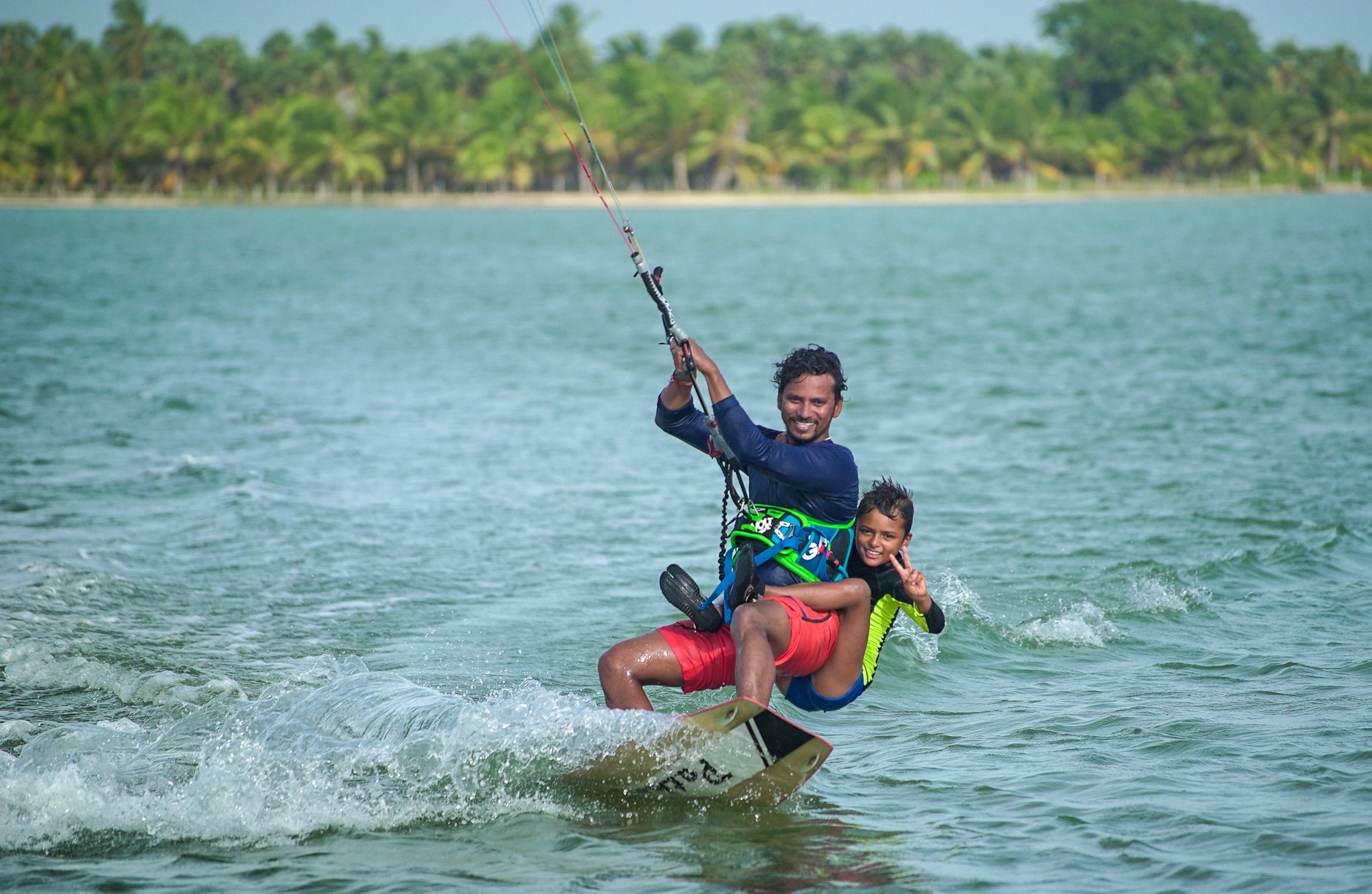 A person windsurfing on a calm sea with a clear sky and a beach with palm trees in the background.