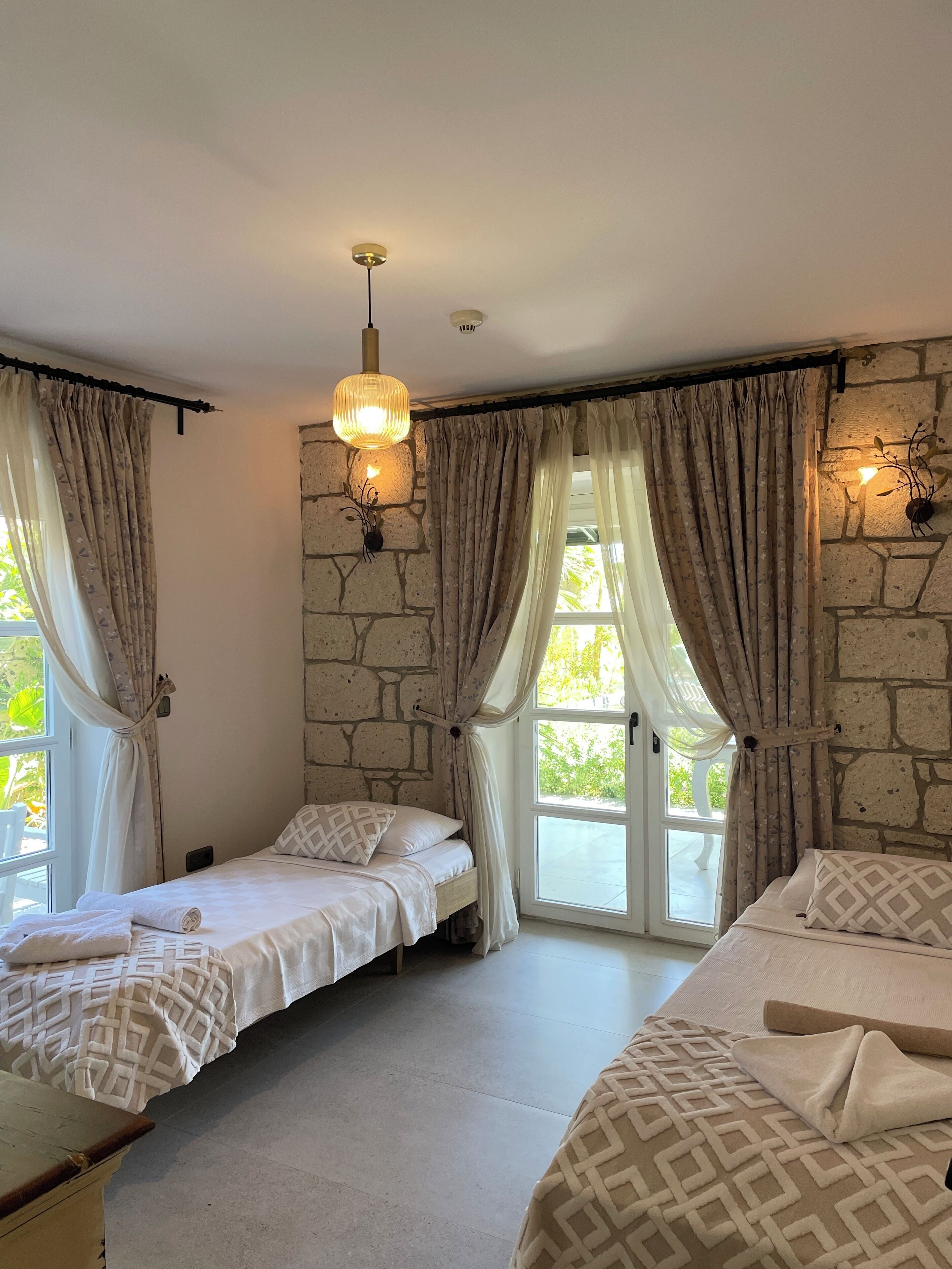 A room with two beds, a stone wall, a hanging light fixture, and a window with curtains.
