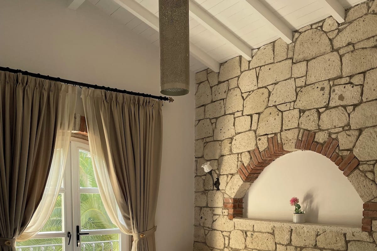 Un dormitorio con pared de piedra, cama con ropa de cama estampada, una mesita y una ventana con cortinas.