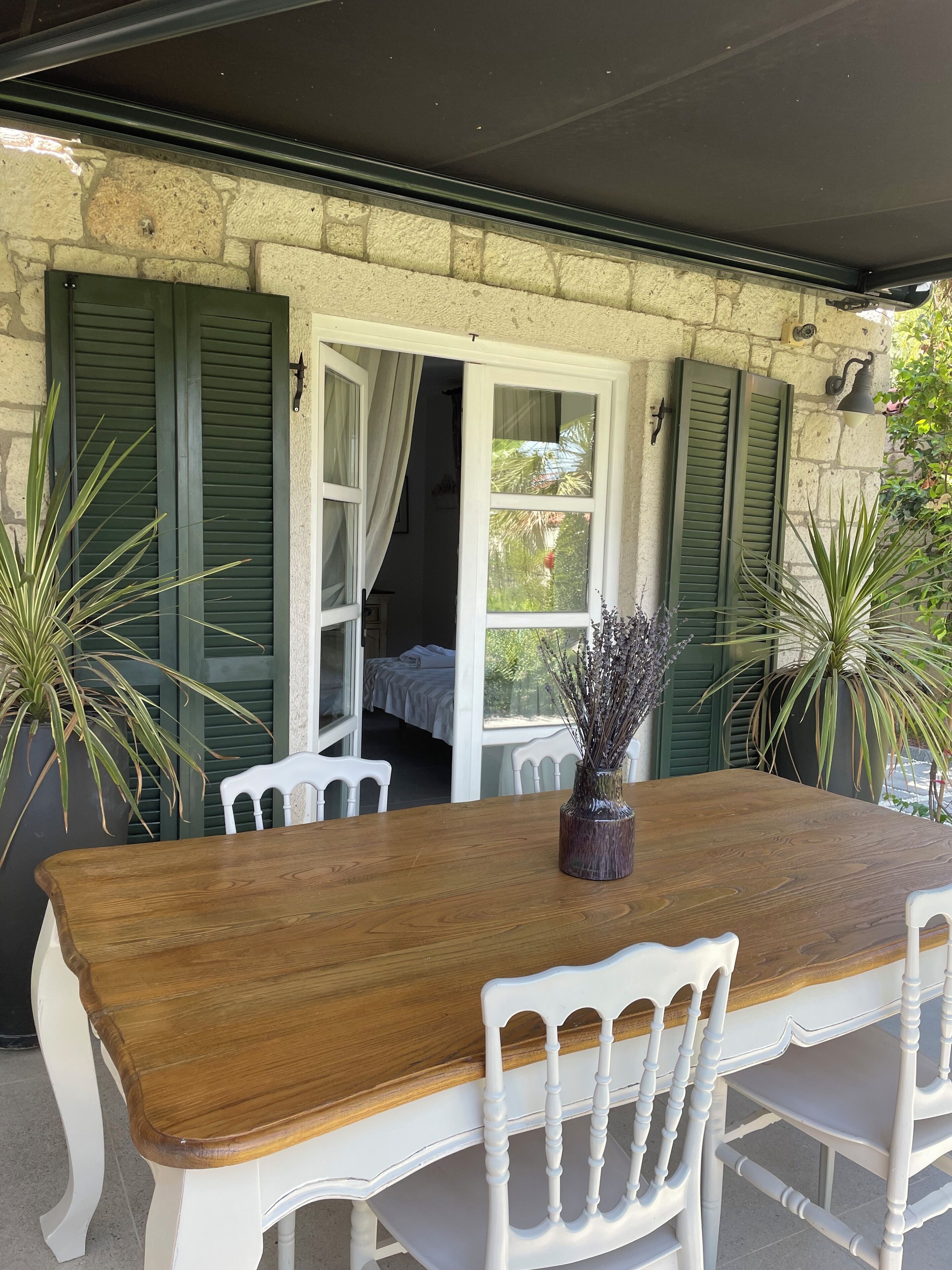 A wooden table with white chairs, green shutters, and potted plants.