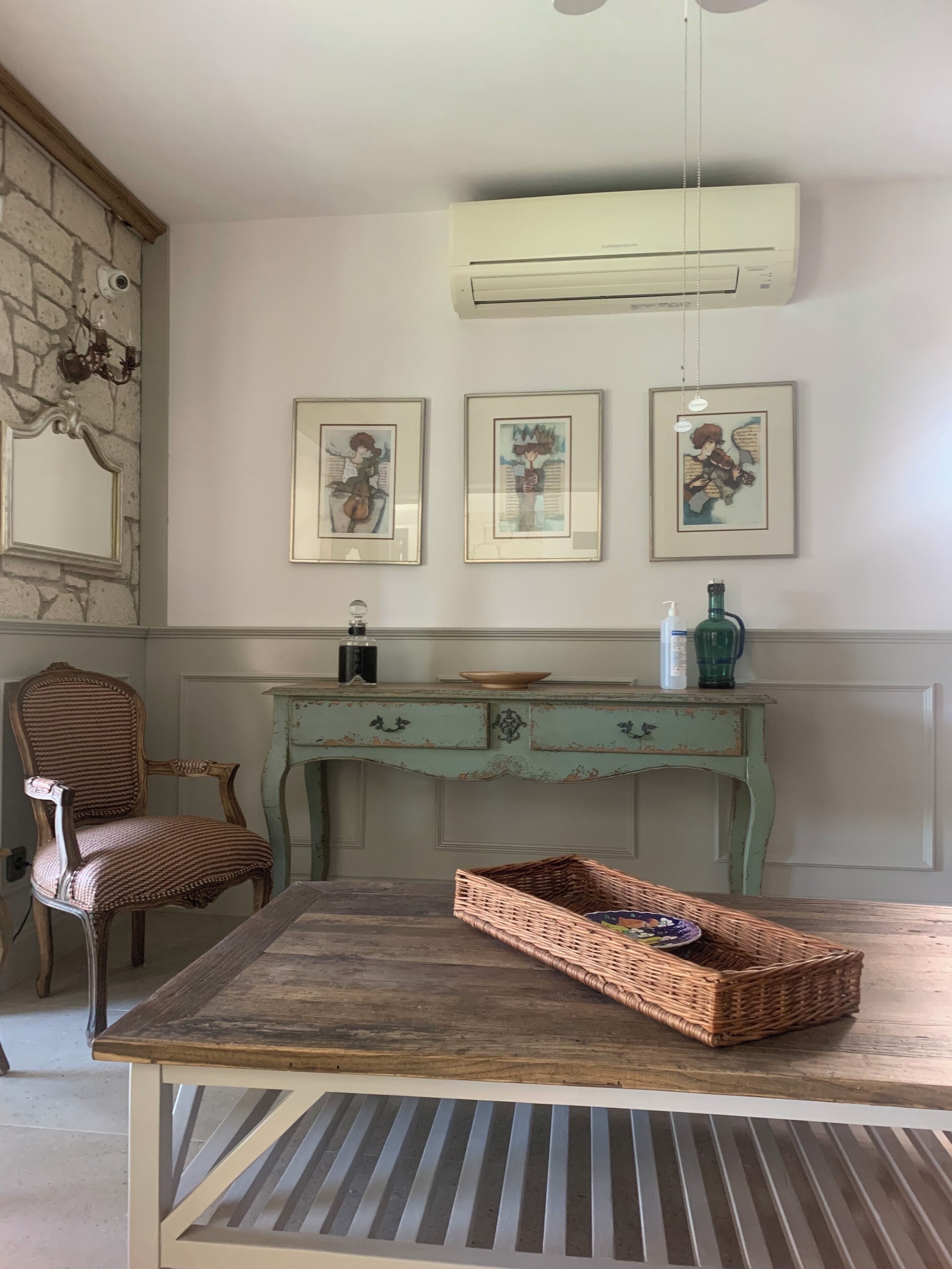 A room with a rustic wooden table, a vintage-style chair, a decorative wall with three framed artworks, a teal-colored dresser, and a ceiling-mounted air conditioning unit.
