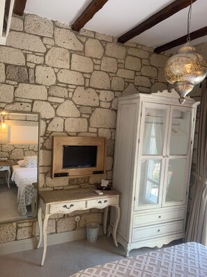 Una habitación con pared de piedra, un escritorio de madera con televisor, un armario blanco y una colcha estampada.