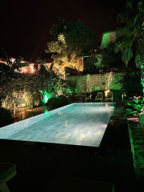 Una piscina con un muro de piedra y follaje, rodeada de sillas y un edificio de piedra al fondo.