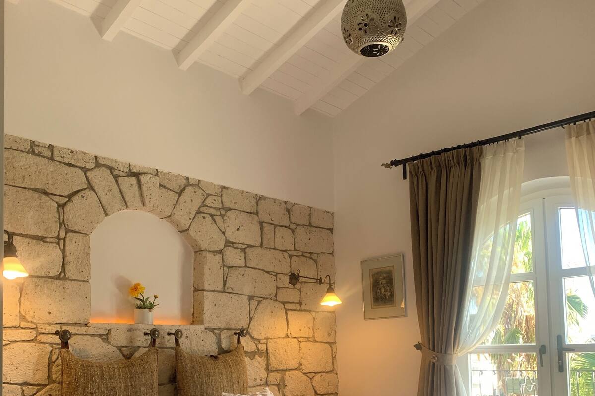 Un dormitorio con pared de piedra, cama con ropa de cama estampada, lámpara colgante y vistas al exterior a través de una ventana.
