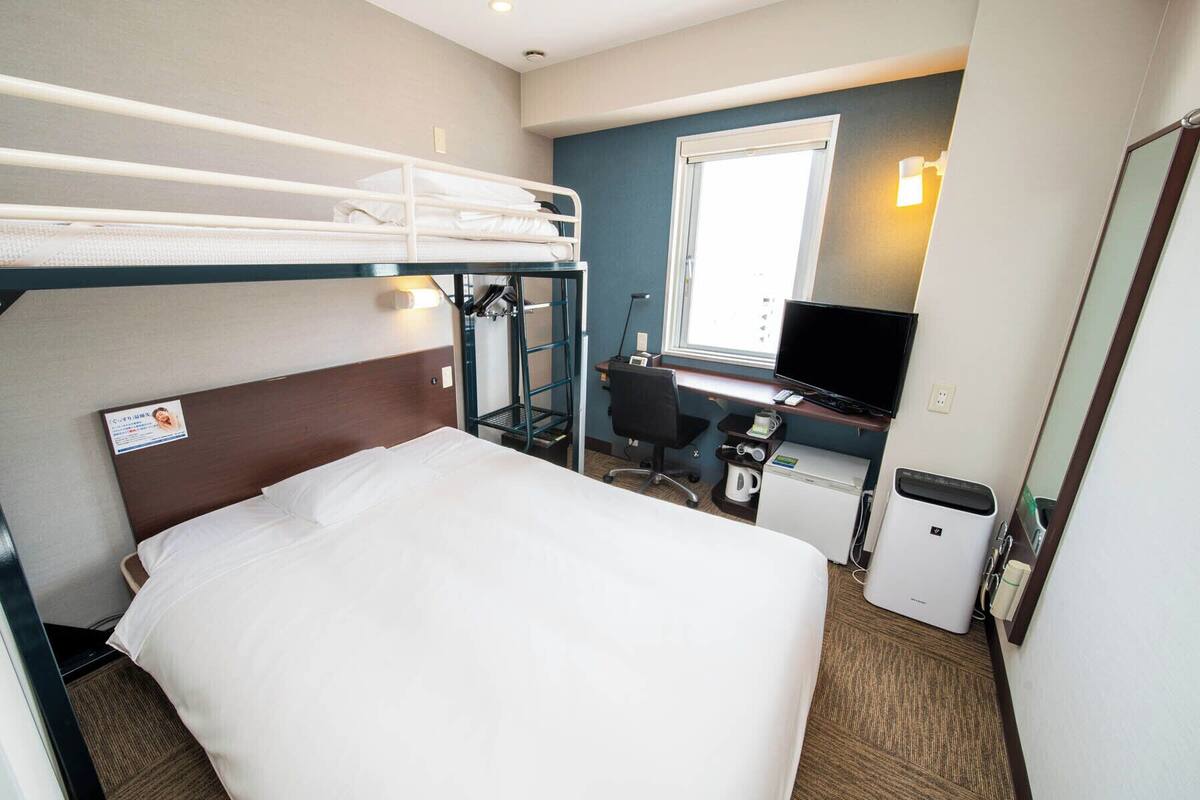 Une chambre d’hôtel avec un lit superposé, un bureau avec un ordinateur, une petite kitchenette et une fenêtre.