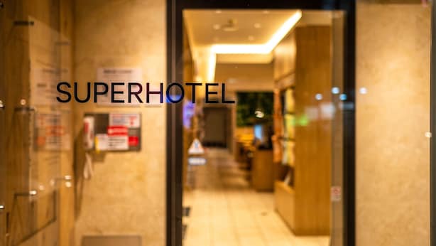 Een hotelingang met een bord waarop 'SUPERHOTEL' staat en een receptiebalie op de achtergrond.