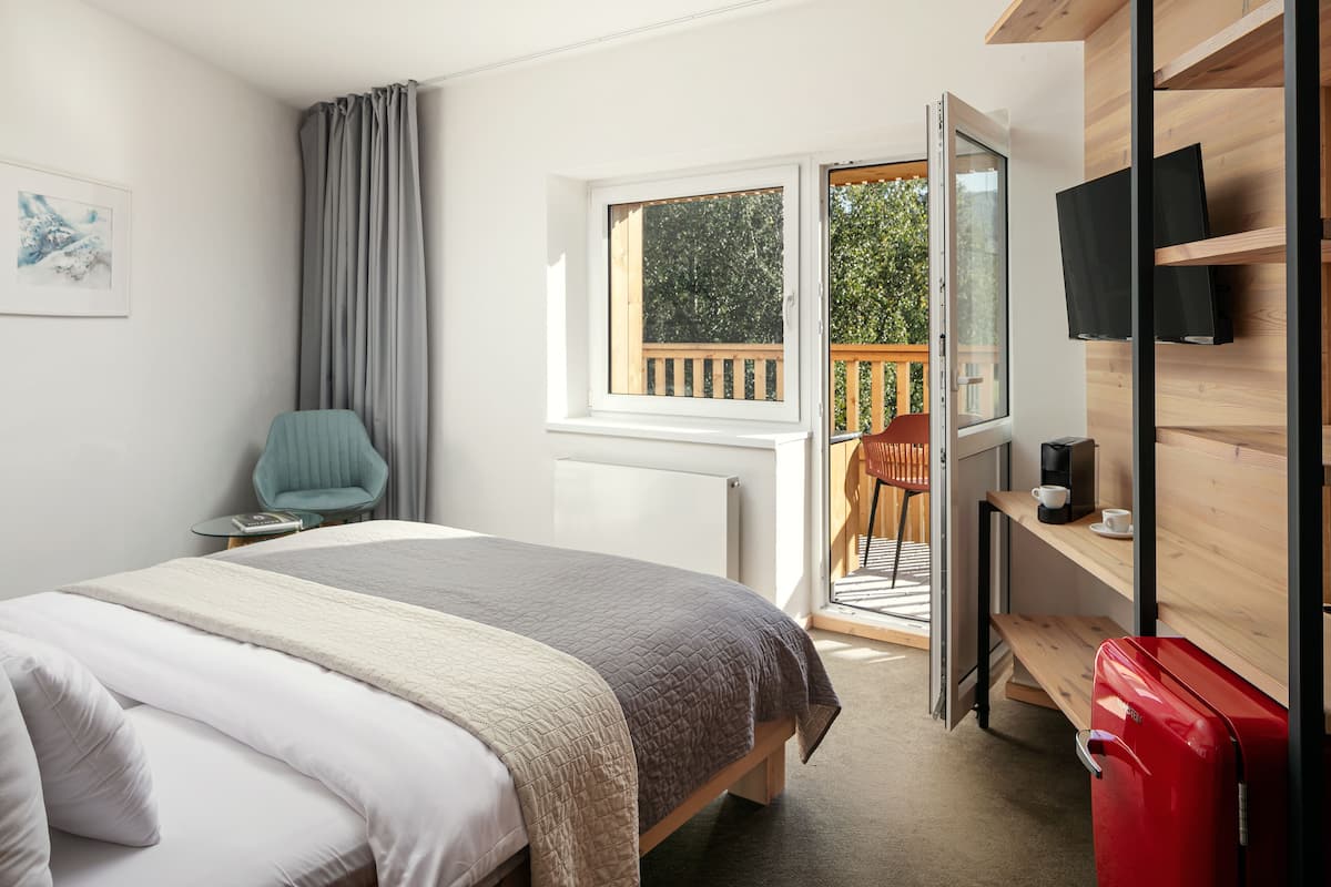 A bedroom with a bed, a chair, a desk, a TV, and a sliding glass door to a balcony.