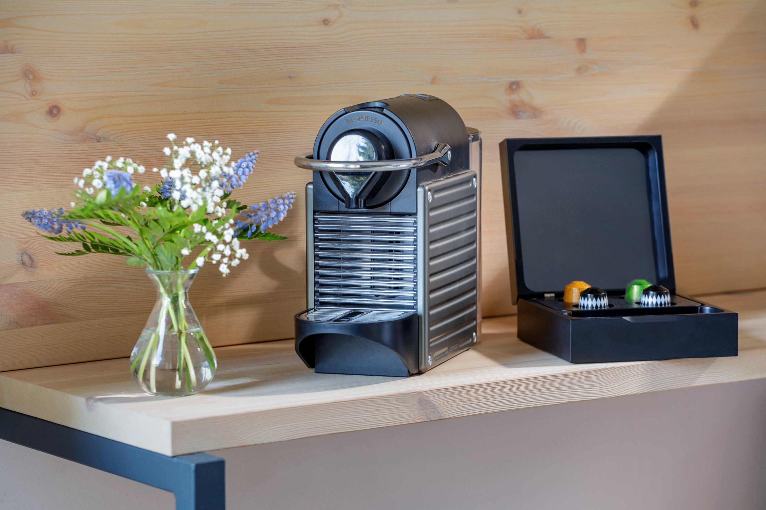 Een koffieapparaat met bekerhouder, een vaas met bloemen en een doos met bestek op een houten oppervlak.