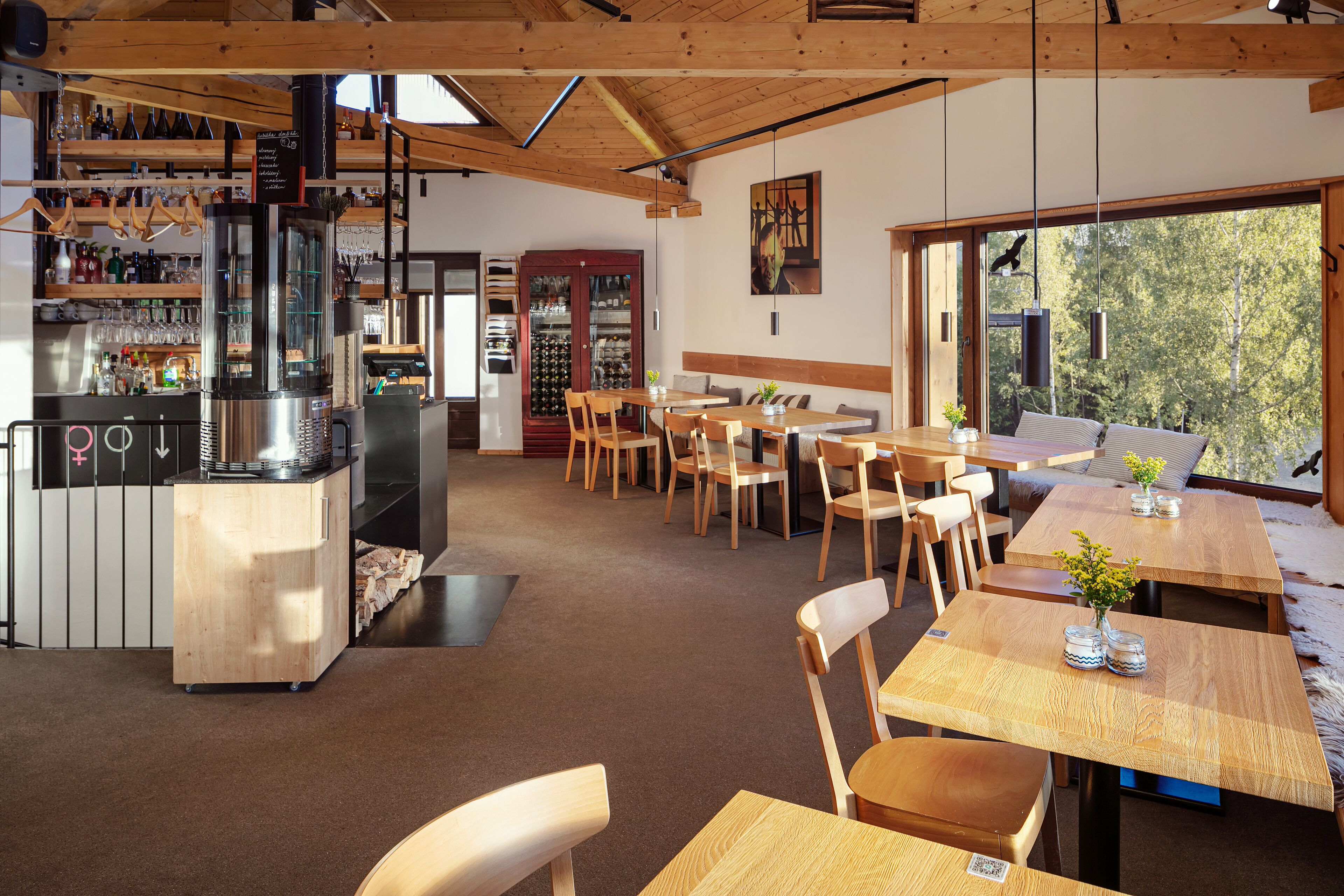 A restaurant with wooden tables and chairs, a bar area with bottles and glasses, and a large window offering a view of trees.