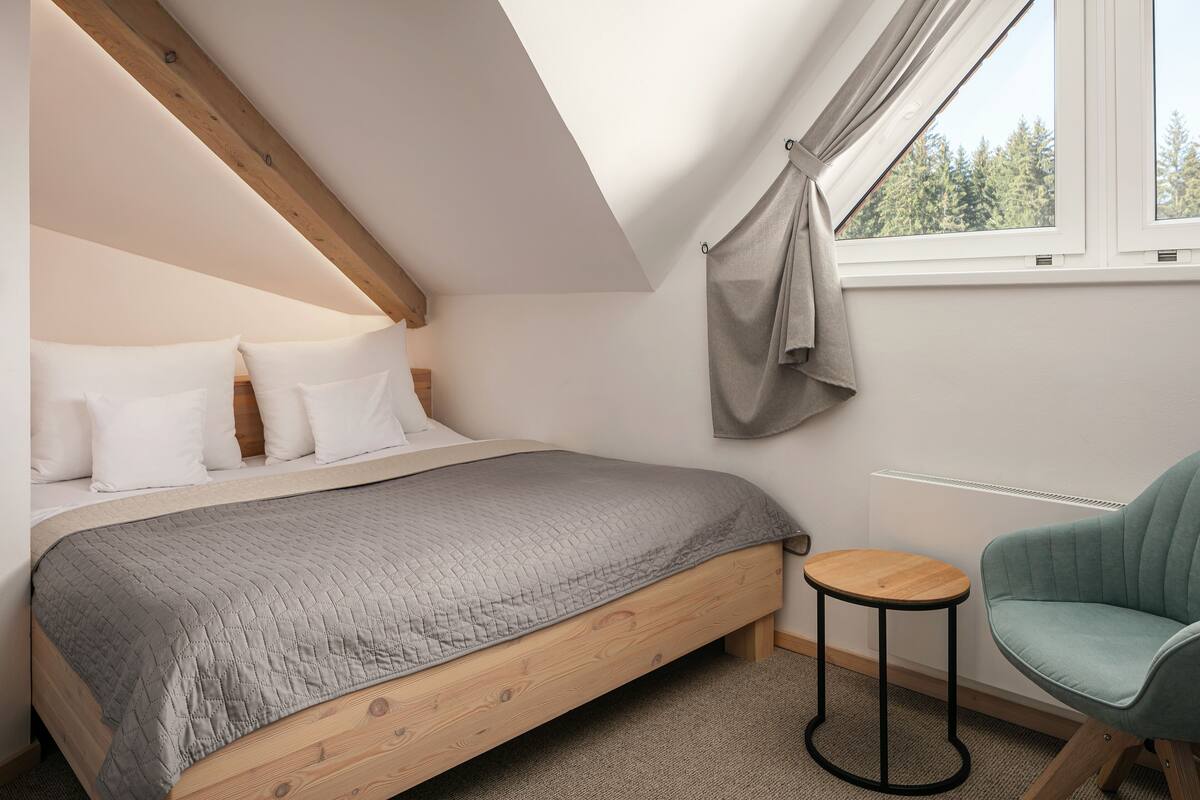 A bedroom with a wooden bed, a grey quilted bedspread, a small round table, and a teal armchair.