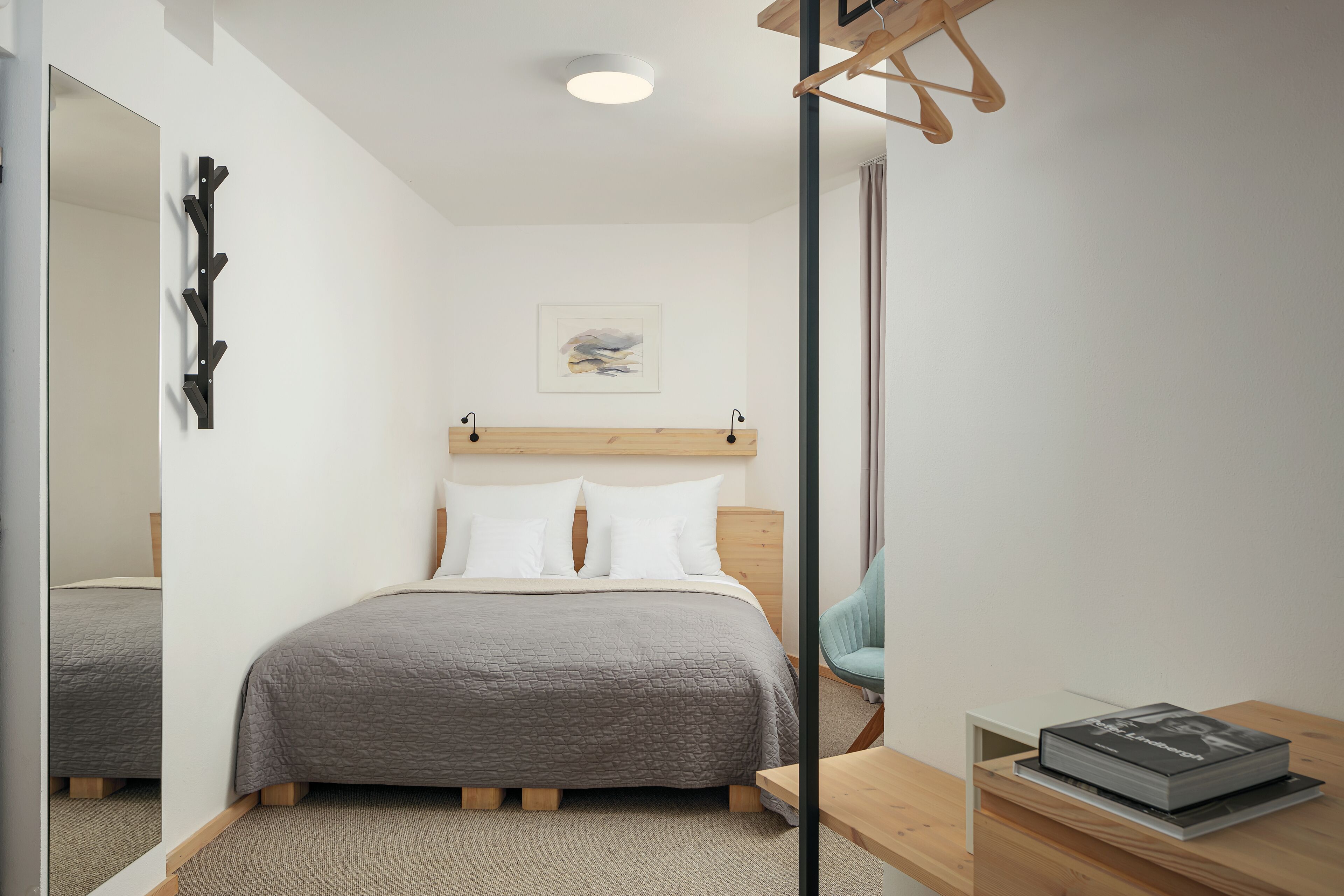 A modern bedroom with a bed, a wooden headboard, a bedside table with a book, a chair, and a mirror.