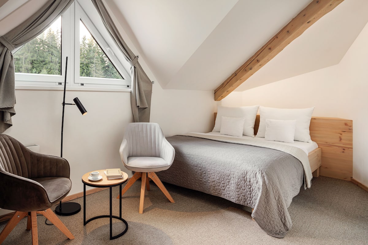 A bedroom with a bed, two chairs, a small table, a floor lamp, and a window with curtains.