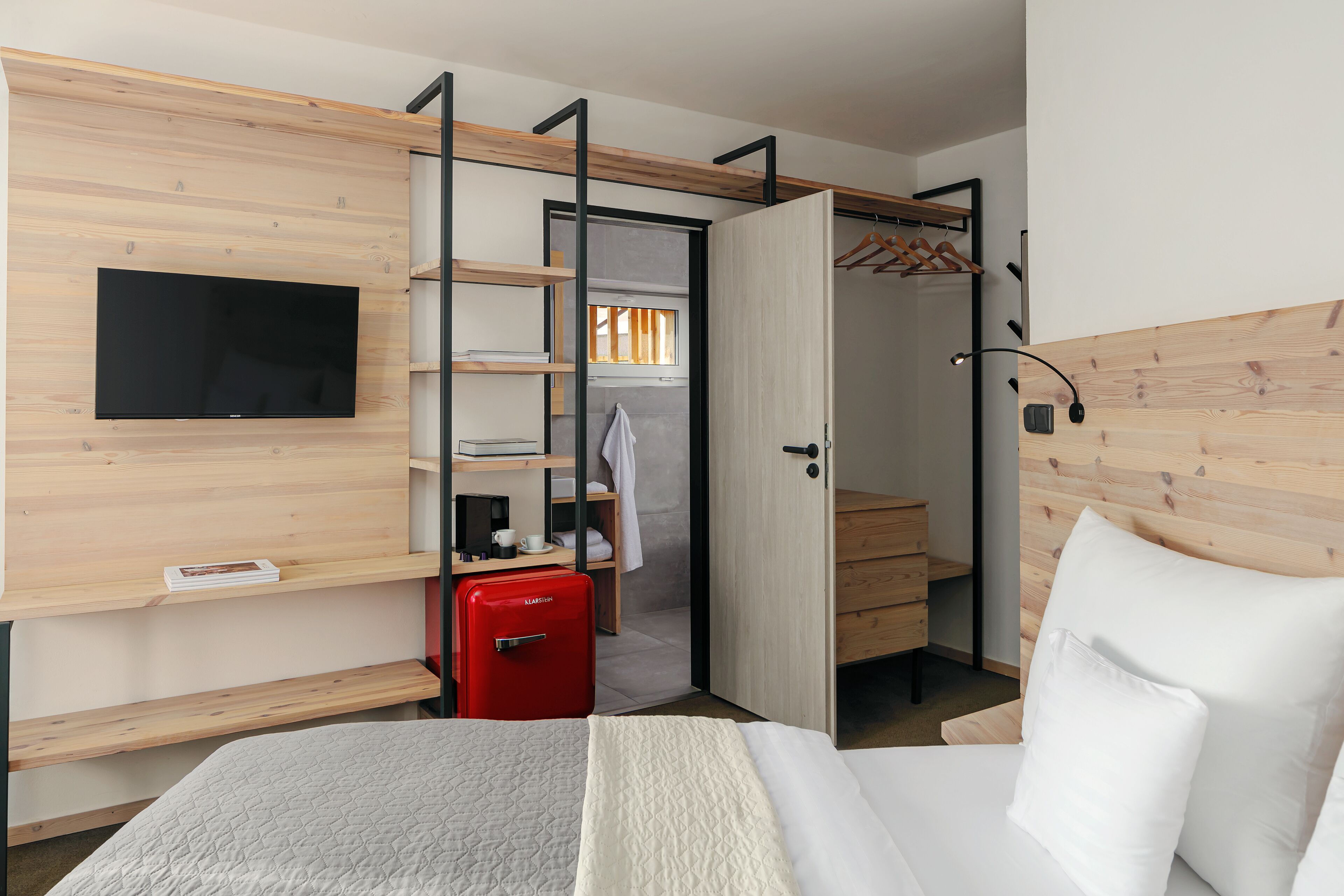 Una habitación de hotel compacta con cama, televisor montado en la pared, una maleta roja y un armario con espejo.