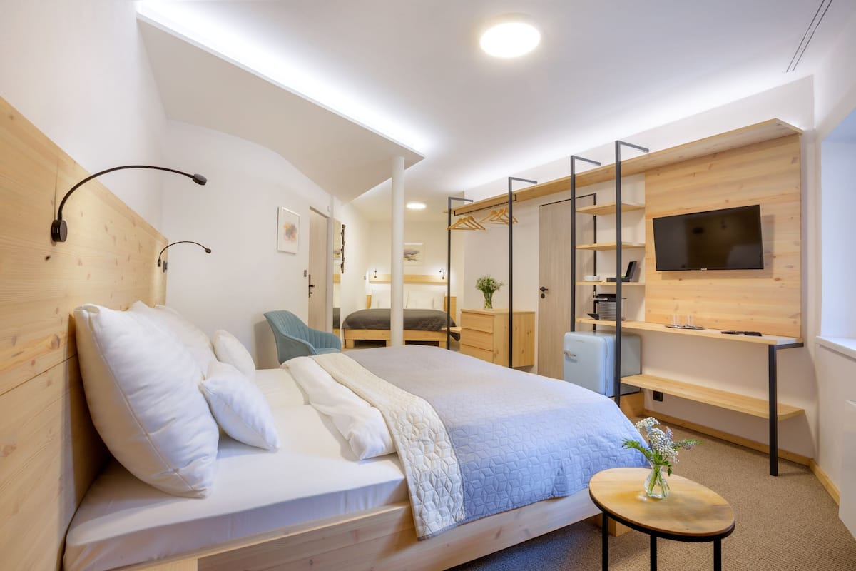 A modern hotel room with a large bed, a TV mounted on the wall, a small table with a vase, and a wooden desk with shelves.
