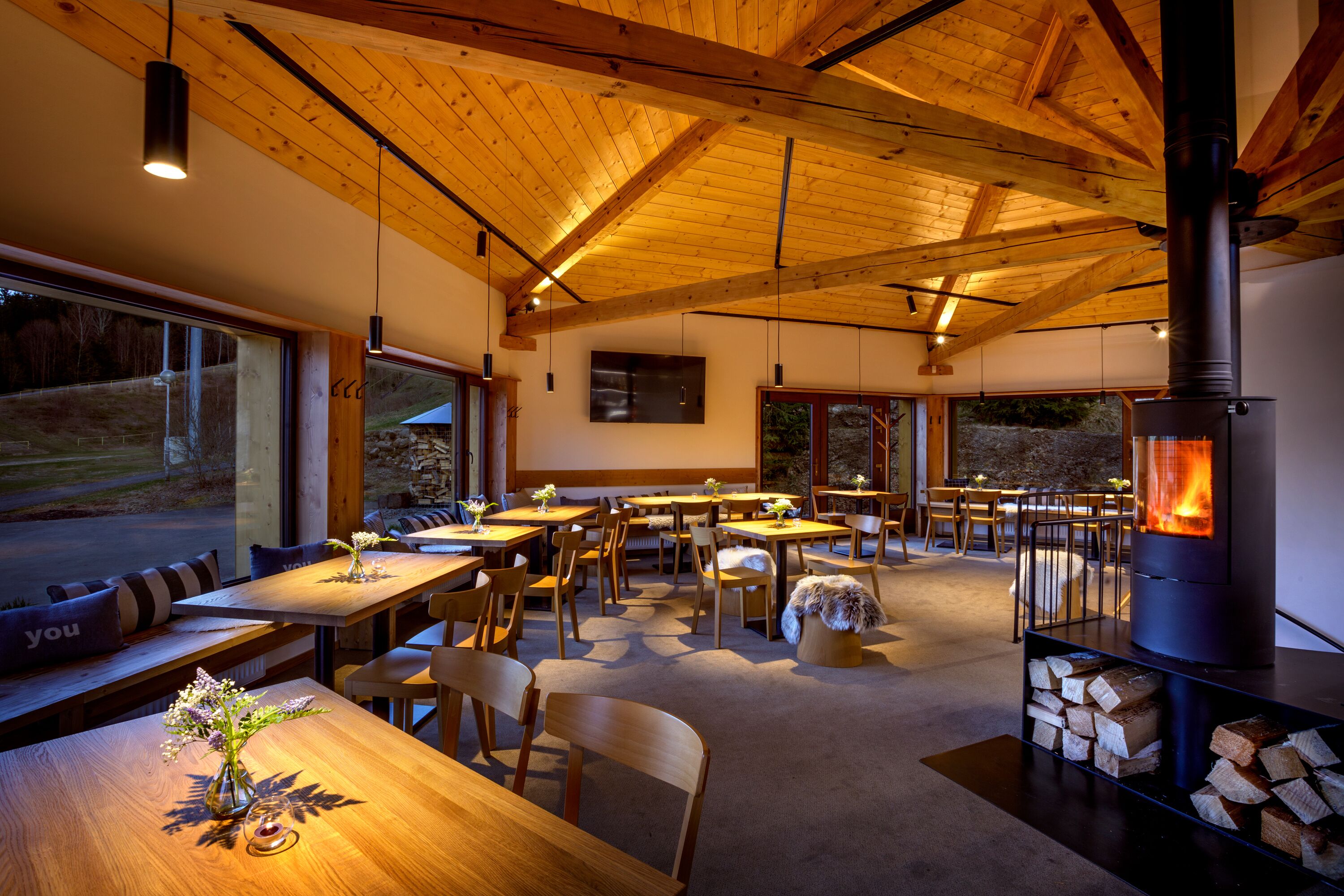 Une salle à manger spacieuse, dotée de tables et de chaises en bois, d’une cheminée et de grandes fenêtres offrant une vue sur l’extérieur.