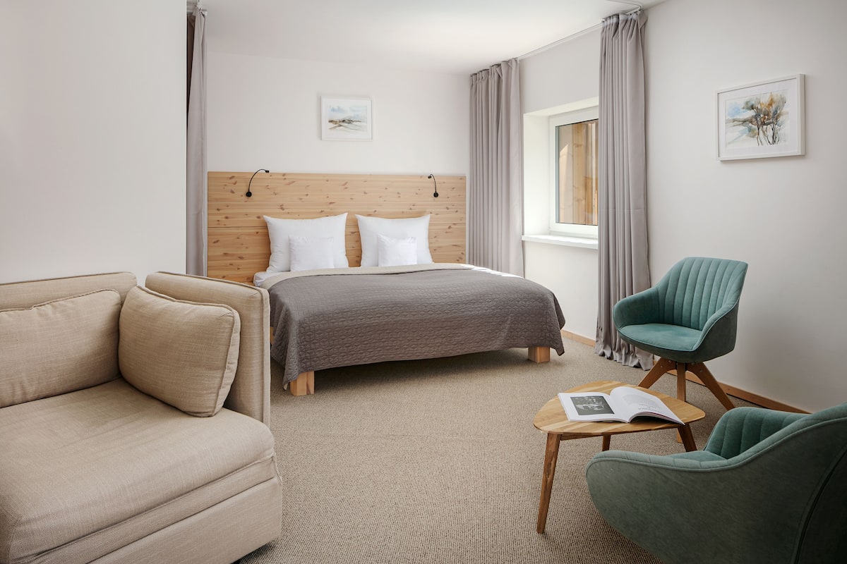 A modern hotel room with a wooden headboard bed, a beige sofa, a teal armchair, and a small wooden side table with an open book.