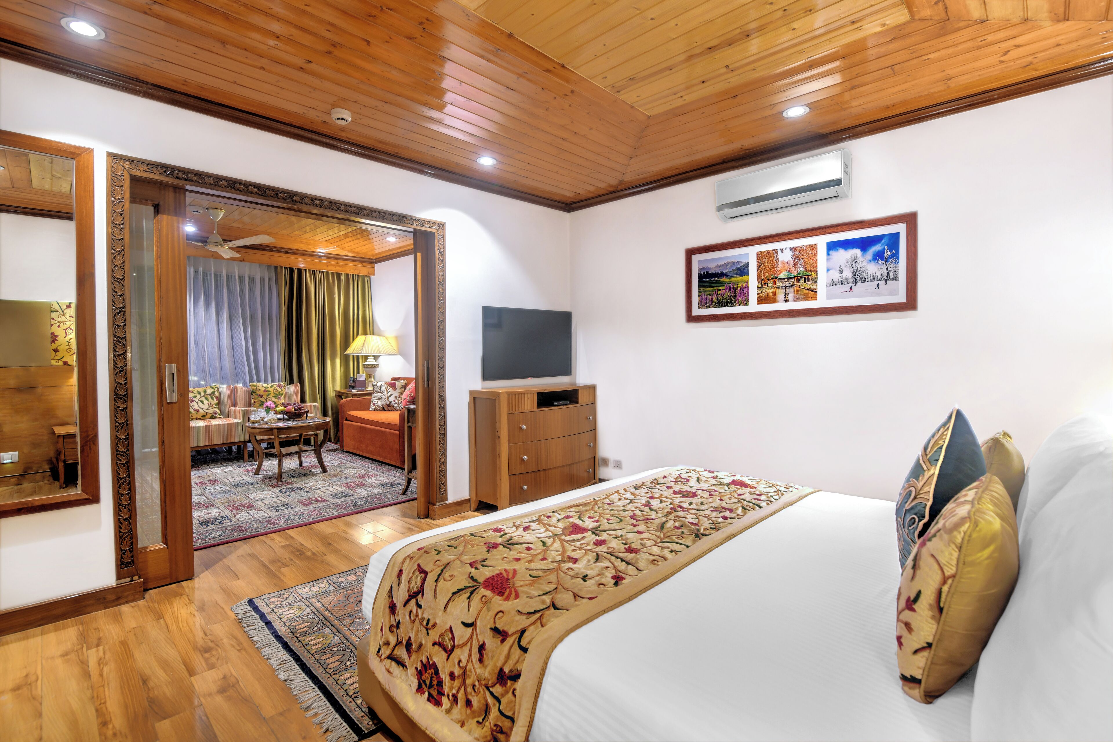 Una habitación de hotel con techo de madera, una cama con ropa de cama floral, un televisor sobre un mueble de madera y una composición de fotos enmarcadas en la pared.