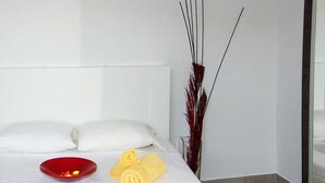 Un letto rifatto con lenzuola bianche, una ciotola rossa e asciugamani arrotolati su un coprimaterasso bianco.