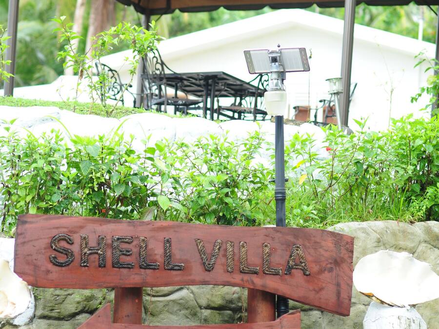Une enseigne en bois portant les inscriptions « SHELL VILLA » et « BIENVENUE », placée devant un mur de pierre surmonté d’un auvent.