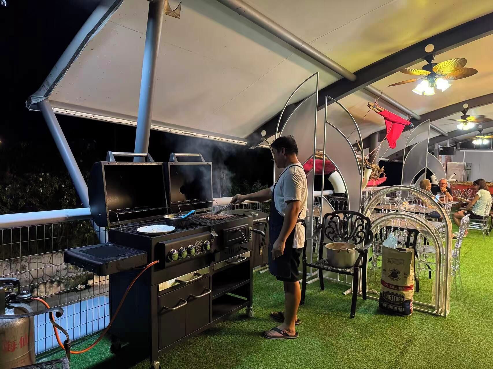Een barbecue-opstelling met een grill, kookgerei en iemand die het eten in de gaten houdt.