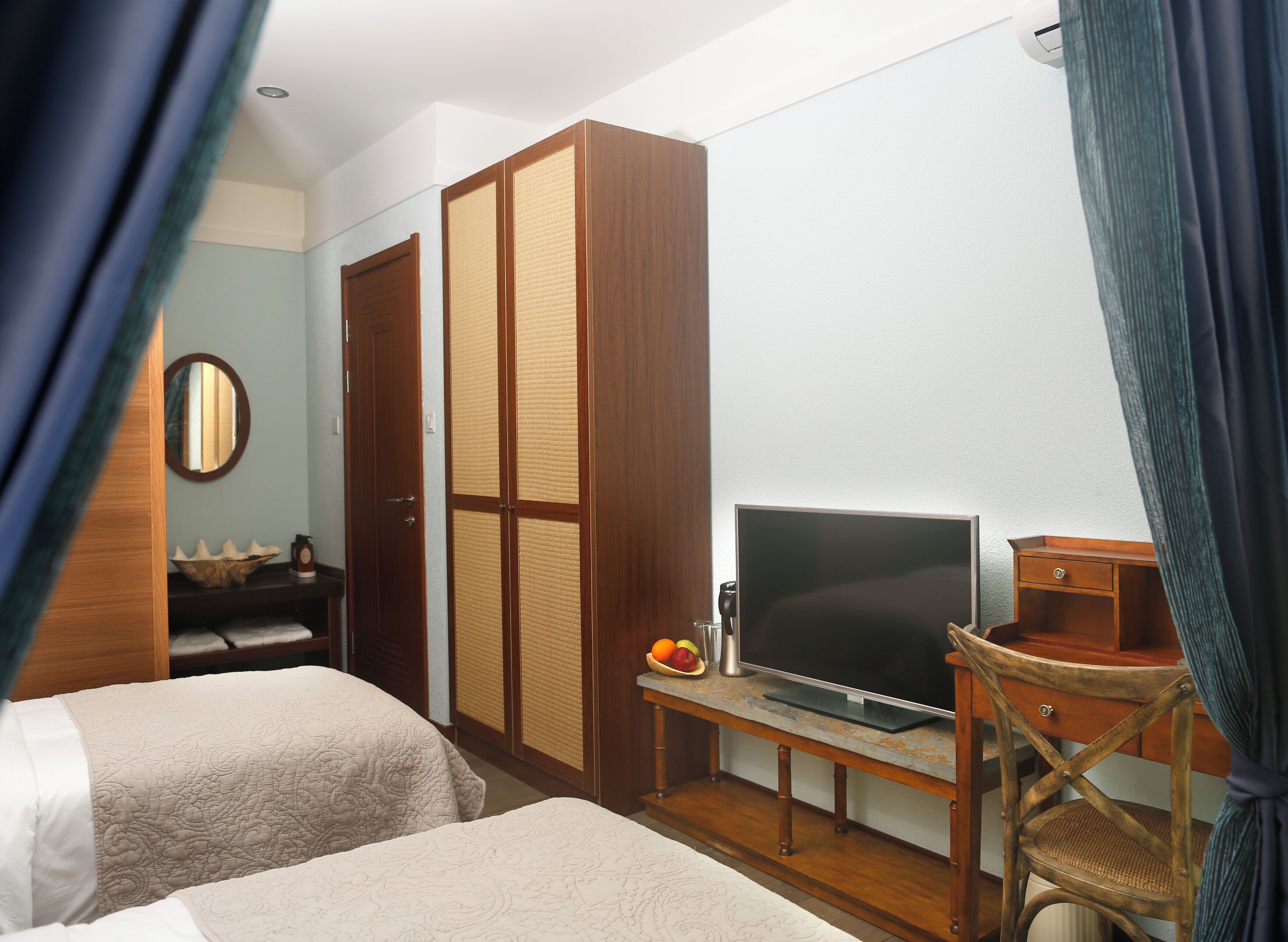 A hotel room with two beds, a TV, a wooden wardrobe, a small table with a fruit bowl, and a chair.