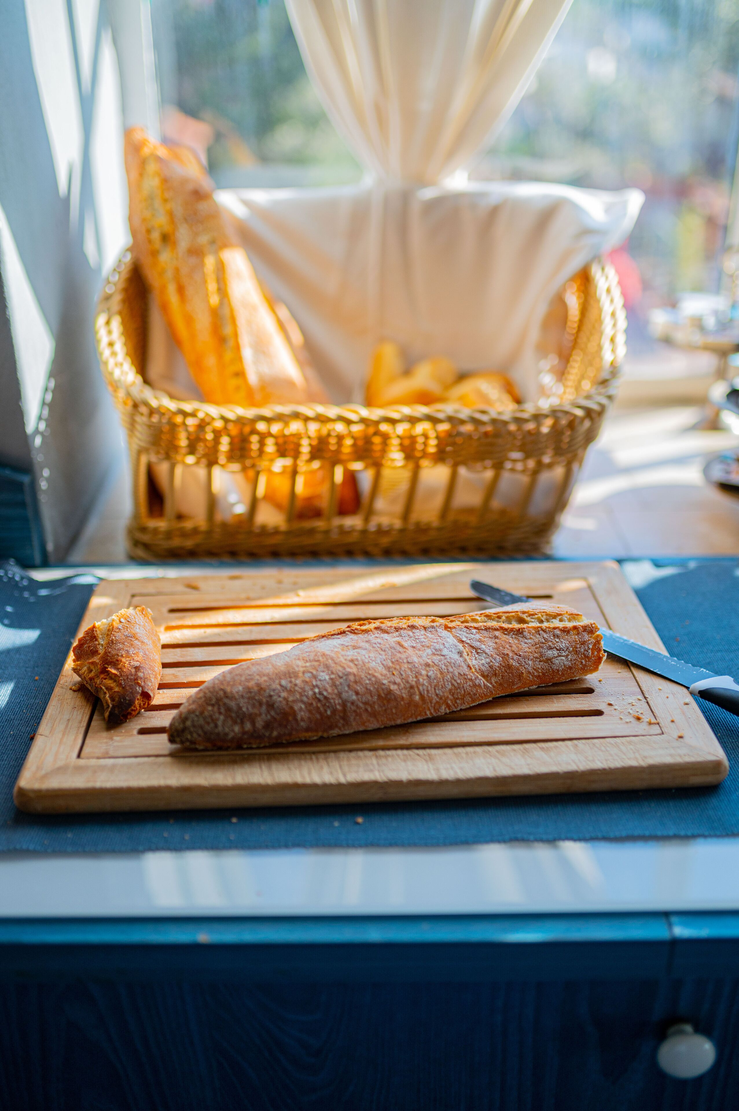 A wooden cutting board with a baguette and a knife on a table near a window with a basket of bread in the background.