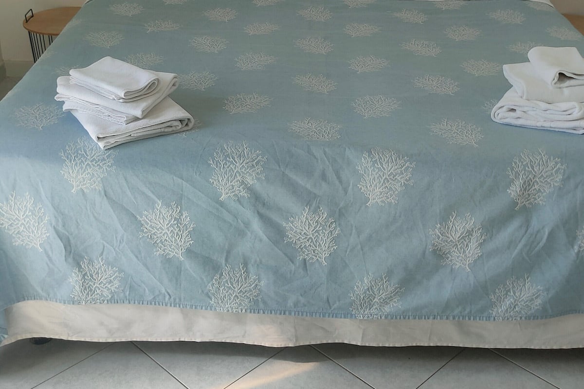 Una cama bien hecha con cubrecama azul y almohadas blancas, una obra de arte enmarcada en la pared y una mesita de noche con una lámpara.