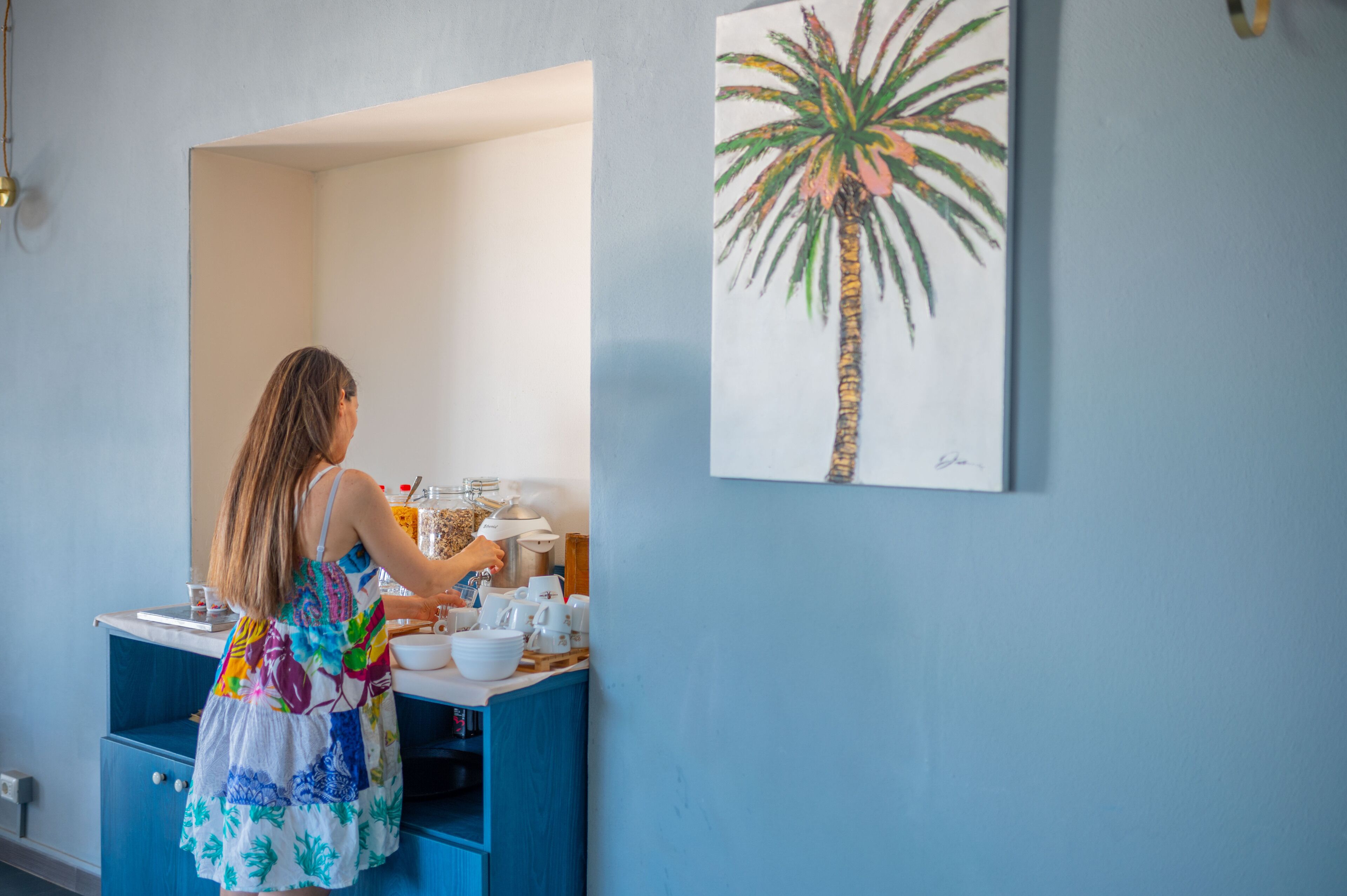 Une personne vêtue d’une robe colorée prépare un repas sur un plan de travail de cuisine, devant un meuble bleu et un tableau représentant un palmier accroché au mur.