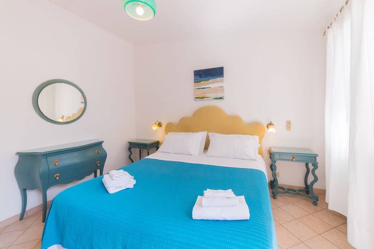 Un dormitorio con una cama, dos mesitas de noche, un tocador, un espejo y un cuadro en la pared.