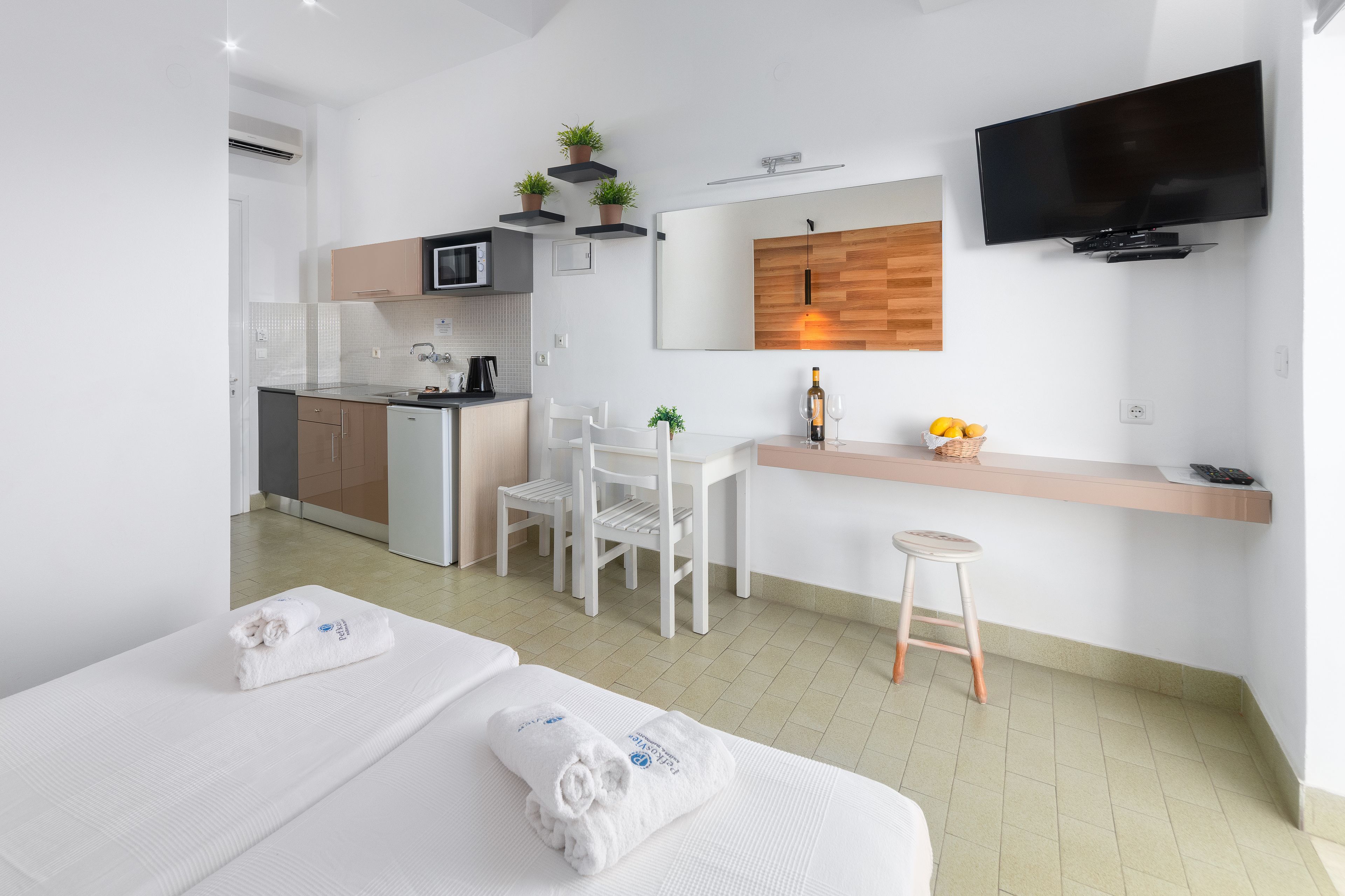 Una habitación de hotel moderna con kitchenette, televisor montado en la pared y un pequeño rincón de estar con una silla y un taburete.