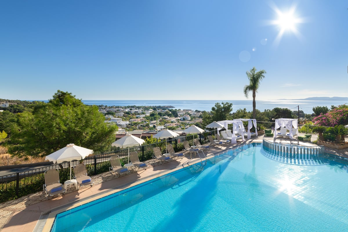 Una piscina con sillas de descanso y sombrillas, rodeada de vegetación y con vistas a un pueblo costero.