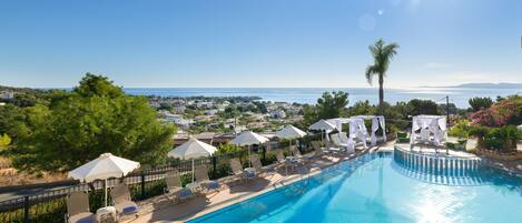 A swimming pool with lounge chairs and umbrellas, surrounded by greenery and overlooking a coastal town.