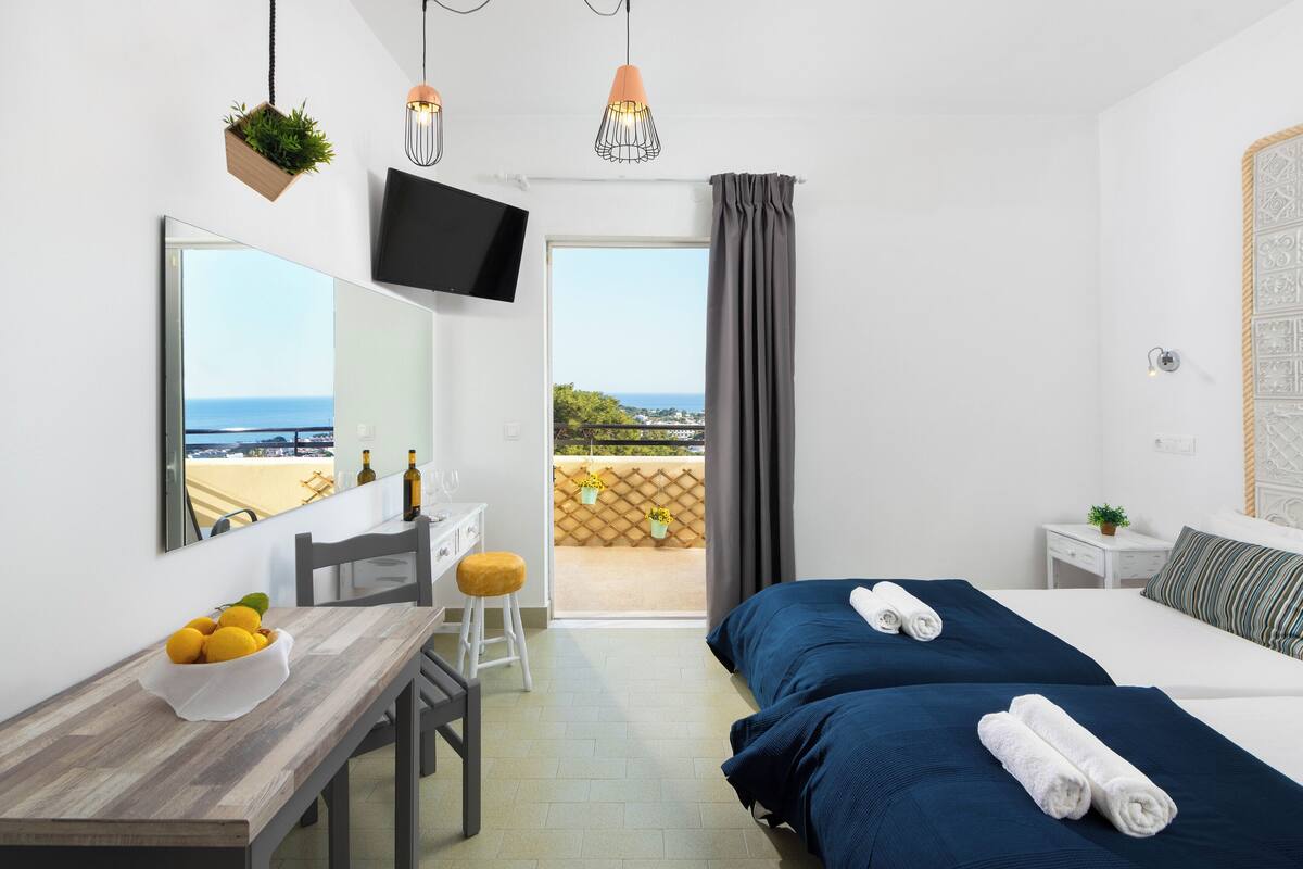 Una habitación de hotel con cama, escritorio con frutero, silla, televisor y balcón con vista al mar.
