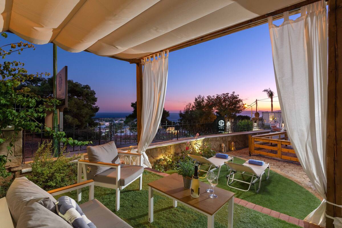 Un patio al aire libre con vista al atardecer, una mesa con una botella y vasos, y una silla de descanso.