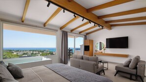 A modern bedroom with a large bed, a sofa, a chair, a TV, and a view of the sea. - Pefkos View Suites & Maisonette (Rhodes)