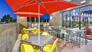 Un patio al aire libre con mesas y sillas, un amplio paraguas rojo y vistas al área del restaurante.