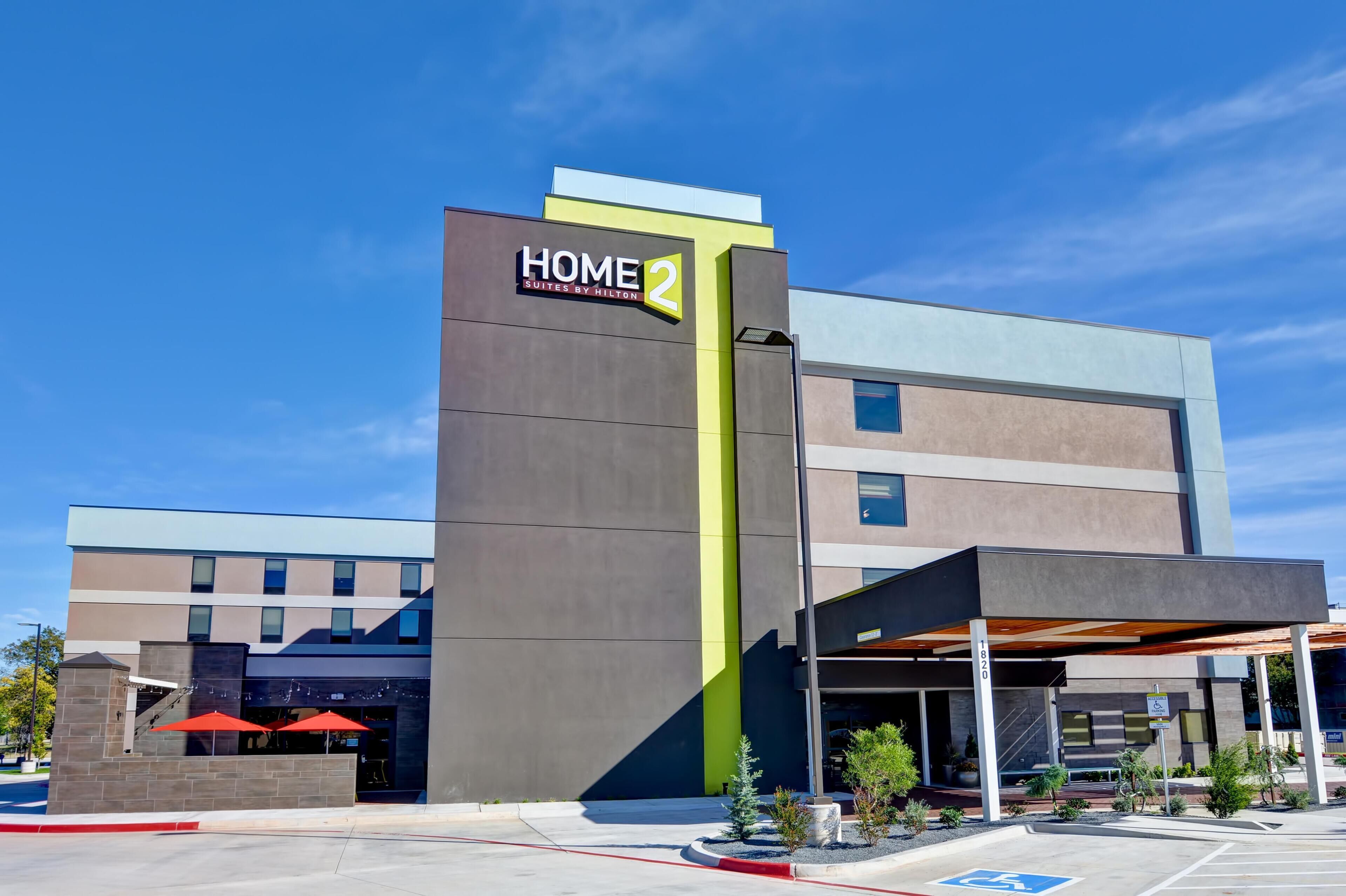 En modern hotellbyggnad med en tydlig skylt som visar ”HOME 2”, en överbyggd entré och en parkeringsplats med en parkeringsplats avsedd för personer med funktionsnedsättning.