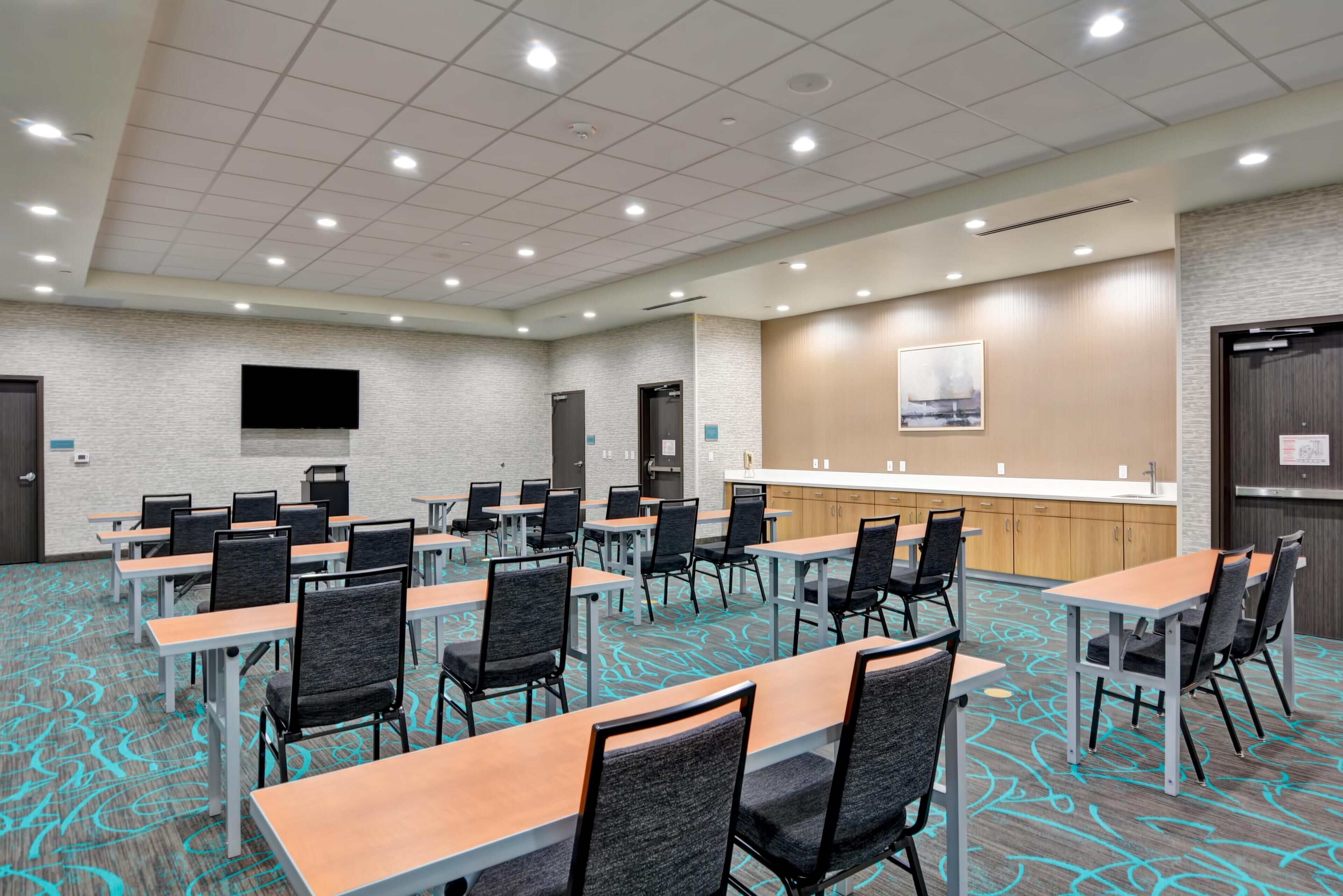 A conference room with tables and chairs, a kitchen area, and a flat-screen TV mounted on the wall.